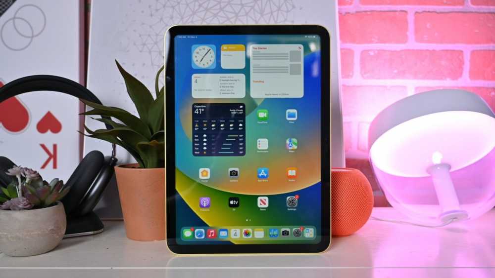 Apple's 10.9-inch iPad sits on a table next to a purple light, AirPods Max, an orange HomePod mini and a plant.