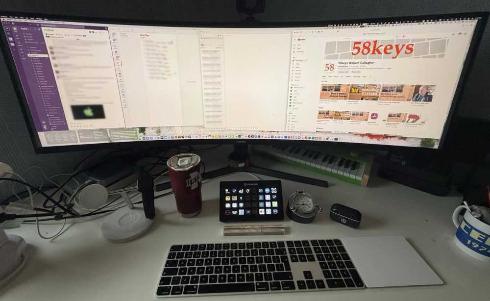 Wide computer monitor displays various applications, with a keyboard, trackpad, tumbler, stream deck, clock, earbuds case, and mug on the desk below.