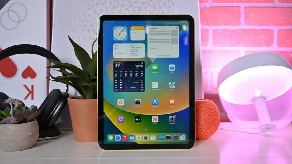 iPad 10代在桌面上显示应用程序和小部件,旁边有两个盆栽、AirPods Max耳机、HomePod mini和一盏发光的灯。