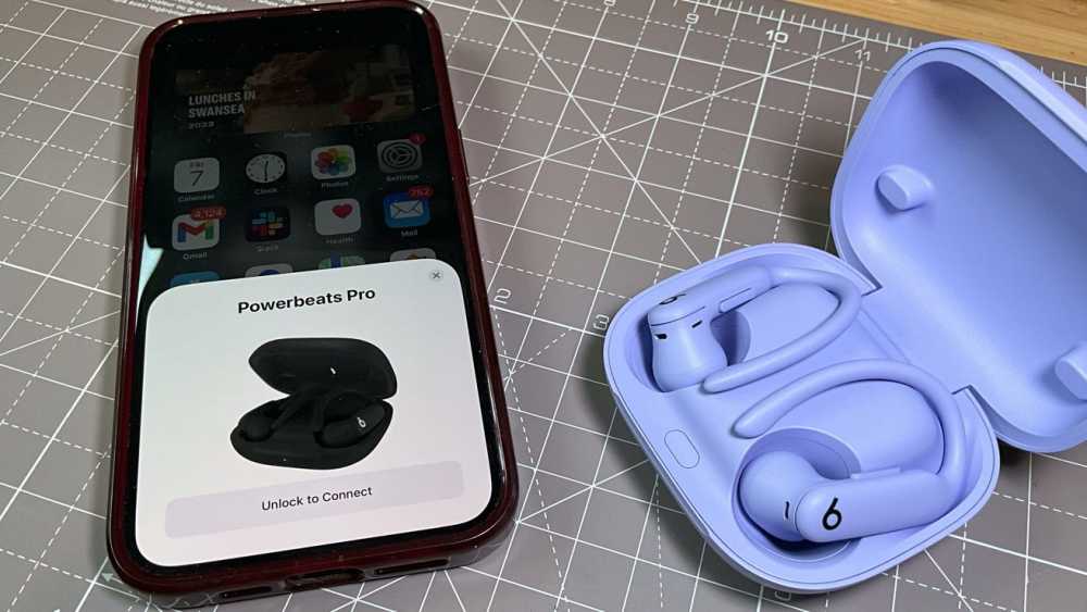一部智能手机显示 Powerbeats Pro 连接通知,旁边是一只打开的薰衣草色充电盒,内有紫色无线耳塞,放在网格图案的表面上。