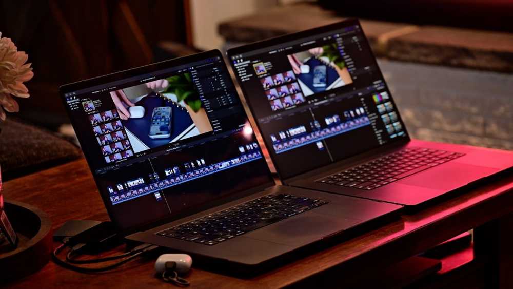 两台 16 英寸 MacBook Pro 笔记本电脑在木制桌子上显示视频编辑软件。附近有一个小盒子和白色花朵的部分视图。背景光线温暖。