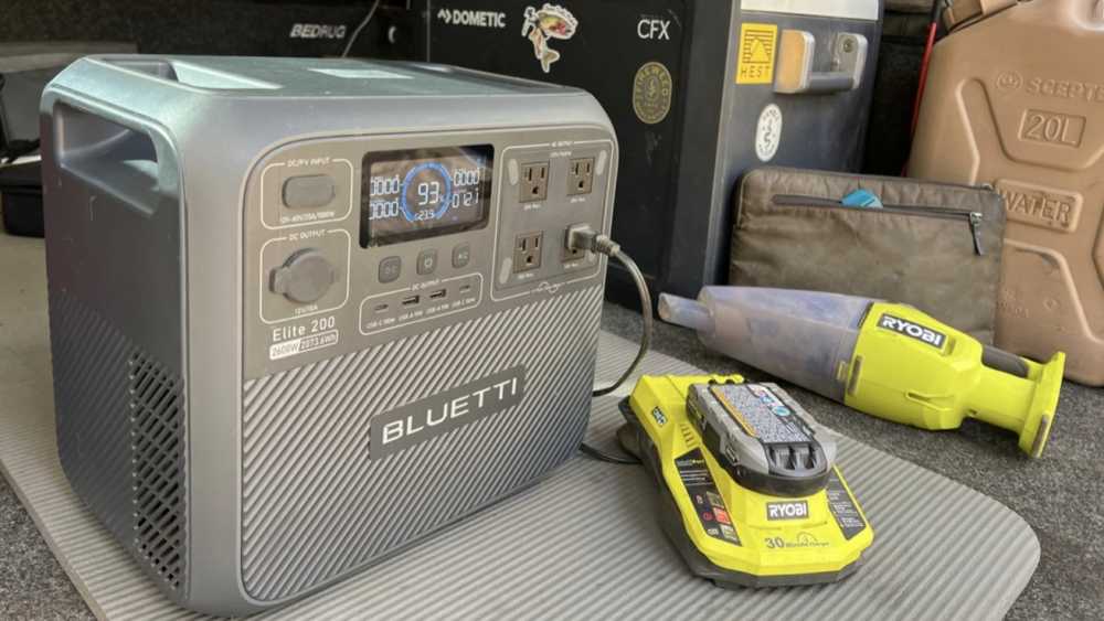 Portable power station with digital display, multiple sockets, and ports; nearby are a charging dock, green handheld vacuum, and a large water container in a utility setting.
