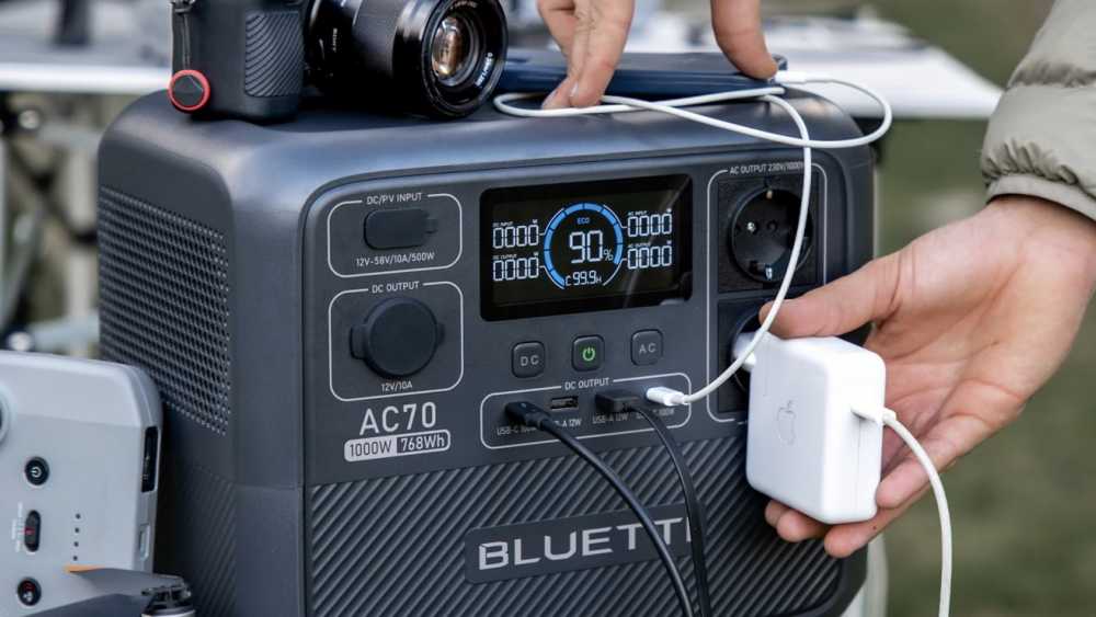 Portable power station with multiple ports and a digital display showing 90% charge. A hand plugs in a charger, and a camera sits on top.