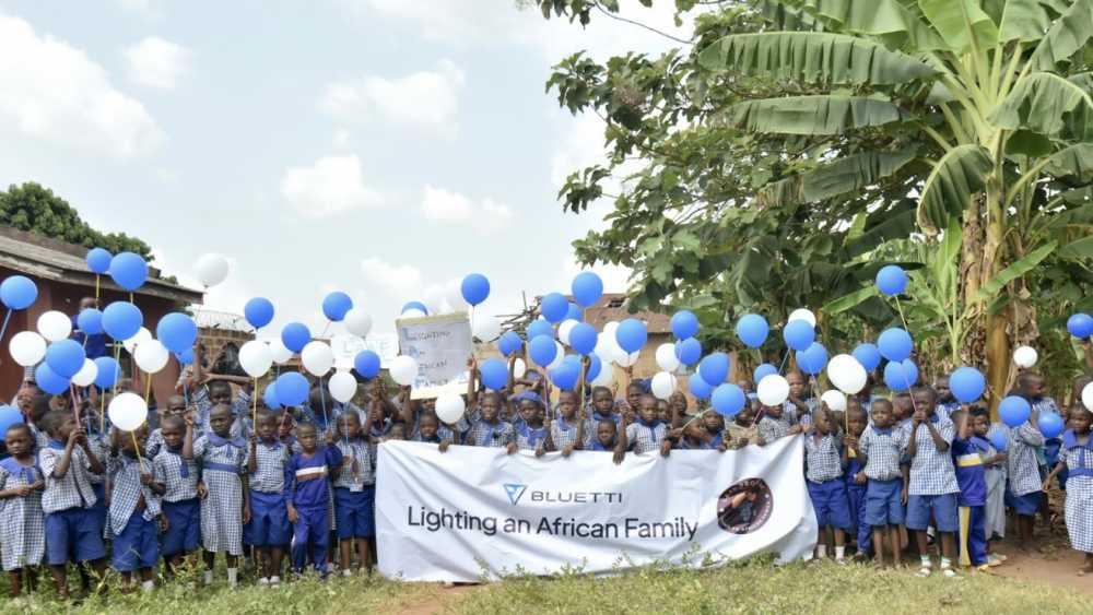 Children in blue-and-white uniforms hold blue and white balloons, standing behind a banner that reads 'Lighting an African Family,' with trees in the background.
