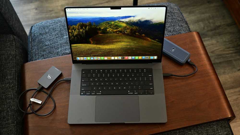 MacBook Pro on wooden table displaying a landscape wallpaper, with two external drives connected, surrounded by gray fabric furniture.