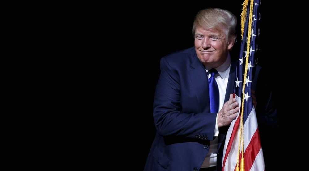一位身穿西装的男士面带微笑,拥抱着一面深色背景下的美国国旗。