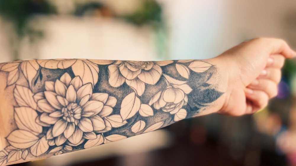 Forearm with detailed black and gray tattoo featuring flowers and leaves, set against a blurred background.