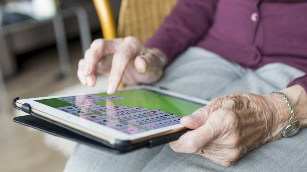 Elderly person playing a card game on a tablet, using fingers to interact with the screen, while seated and wearing a bracelet and purple cardigan.