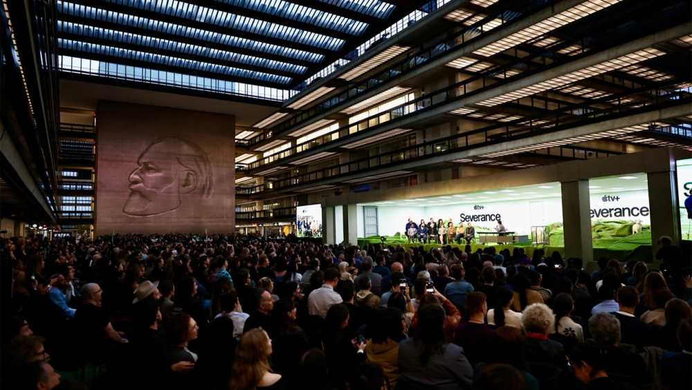 Crowd watching a panel discussion in a large modern atrium, featuring a carved wall portrait. Brightly lit stage with people seated, promotional screens display Severance and Apple TV+ logos.