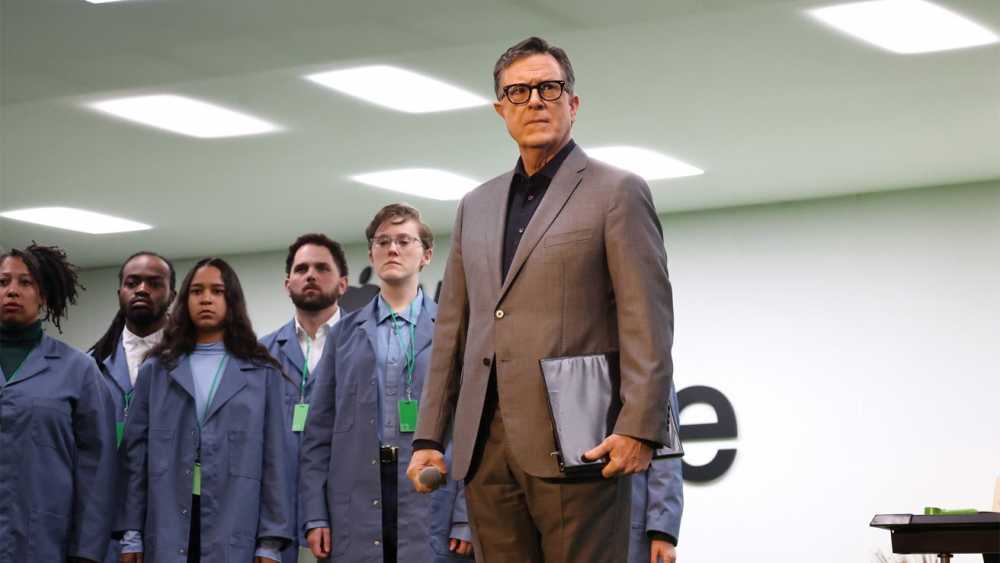A man in a suit holds a folder and microphone, standing with five people in blue coats, under bright ceiling lights.