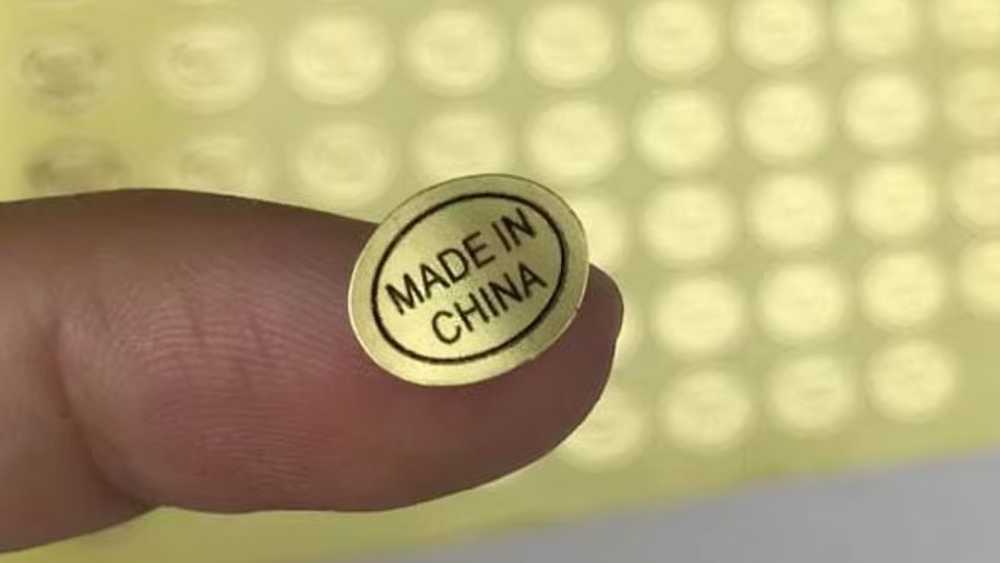 一张小小的金色贴纸在指尖上显示“中国制造”，背景中是模糊的类似贴纸图案。