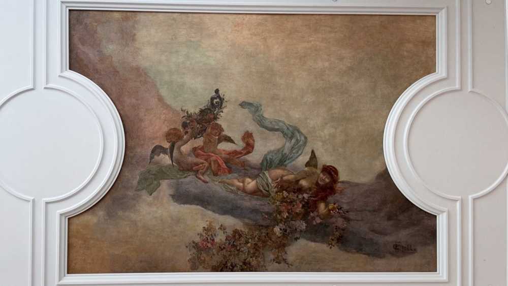 小天使的画作，配有翅膀和一个躺卧的人物，周围环绕着鲜花和飘逸的织物，设置在一个装饰性的白色面板中。