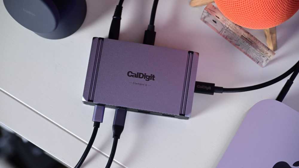 A CalDigit Element Hub with multiple connected wires rests on a light-colored desk, surrounded by electronic devices and accessories.