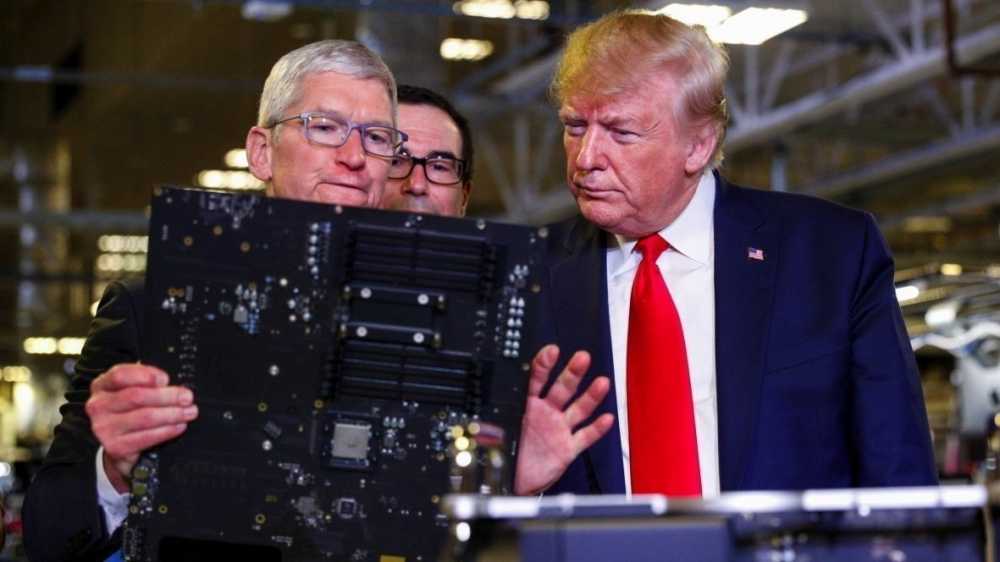 Three men in a factory inspect a large circuit board; one points at it, another wears a red tie, with shelves and lights in the background.