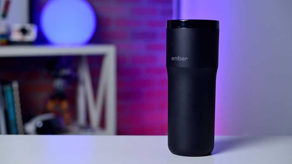 A black travel mug labeled 'ember' on a white table, with a blurred purple and red-lit background featuring shelves and books.