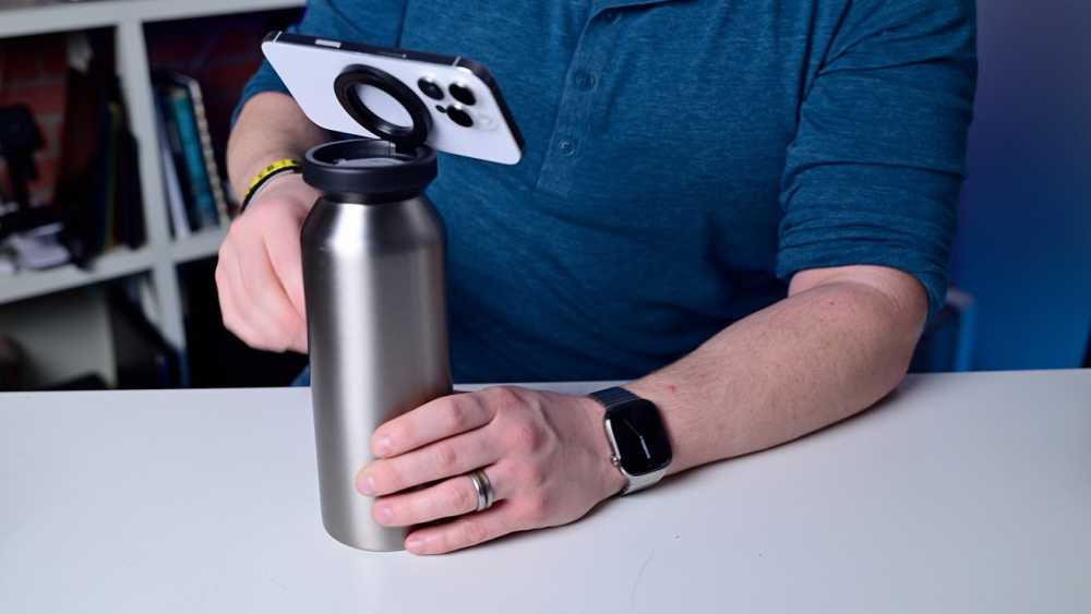 Person wearing a smartwatch holds a metal bottle on a table. A phone is attached to the bottle cap with a ring holder. Books are visible in the background.