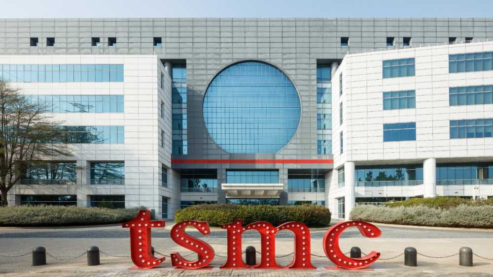 Large modern building with blue-tinted windows, gray circular window feature, and red 'tsmc' letters in front, surrounded by trees and shrubs.