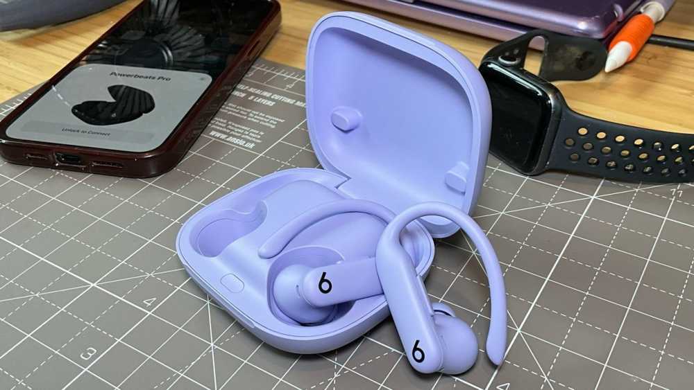 Purple wireless earbuds with ear hooks in an open charging case on a grid mat, with a smartwatch, smartphone, and pens nearby.