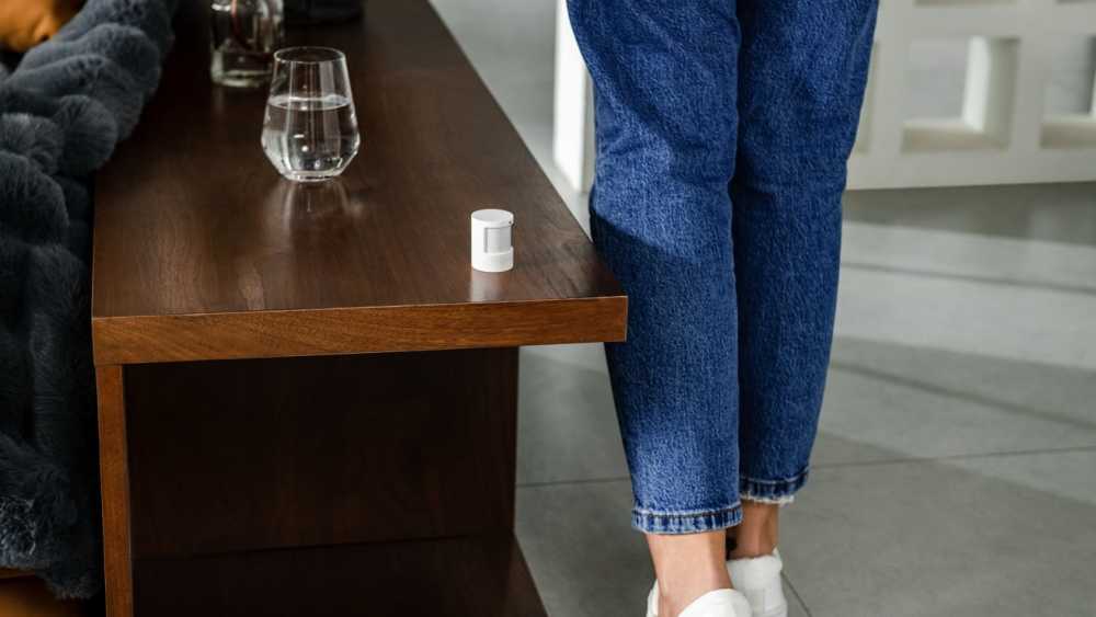 A wooden table with a glass of water and a small white cylindrical object. Person in blue jeans and white shoes stands nearby.