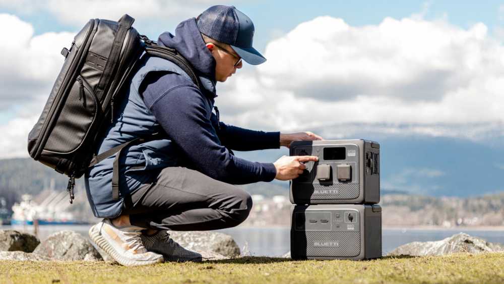 Man squatting outdoors, wearing a cap and backpack, operating two stacked portable power stations, with a mountainous landscape and cloudy sky in the background.