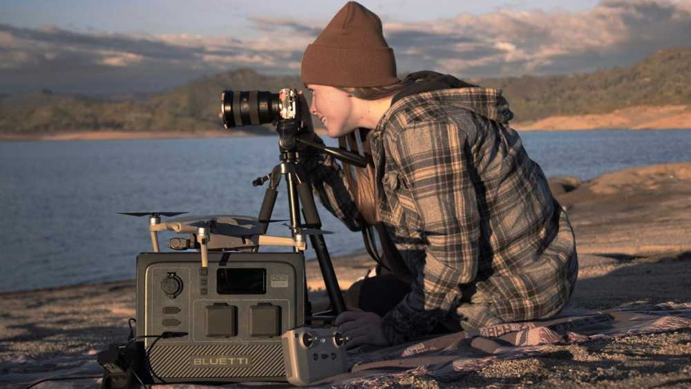 Person in a plaid shirt and beanie photographing a lake landscape with a camera on a tripod, next to a portable power station and a drone controller.