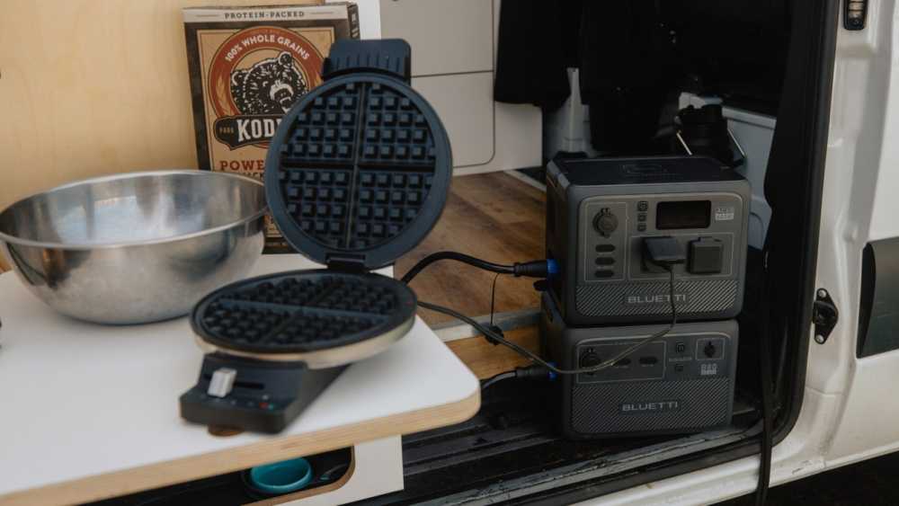 Waffle maker and mixing bowl on a table in a van, with two portable power stations plugged in nearby. Box of pancake mix on the counter.
