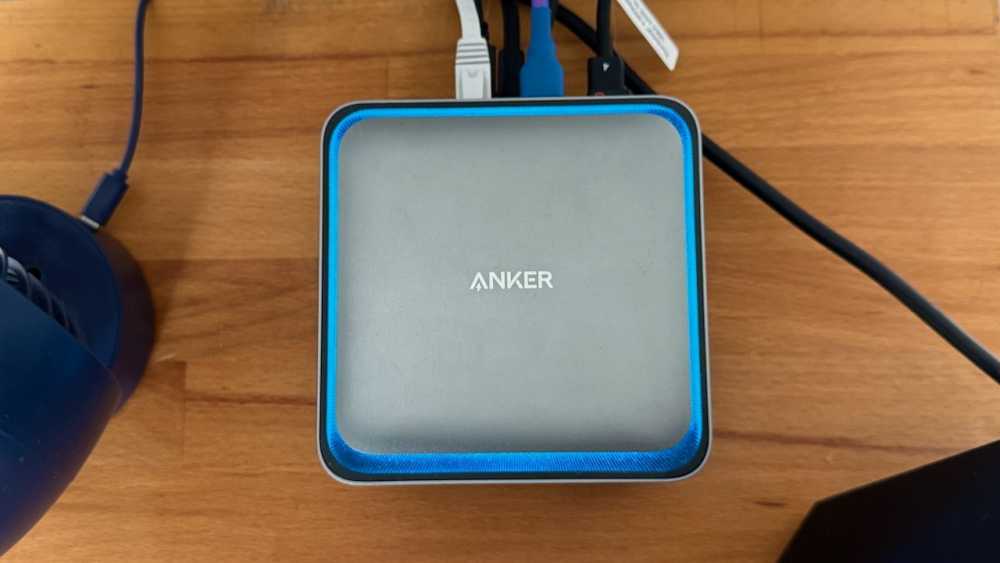 一个小的方形设备，上面标有“Anker”，位于木质表面上，周围连接着电缆，带有蓝色 LED 灯。