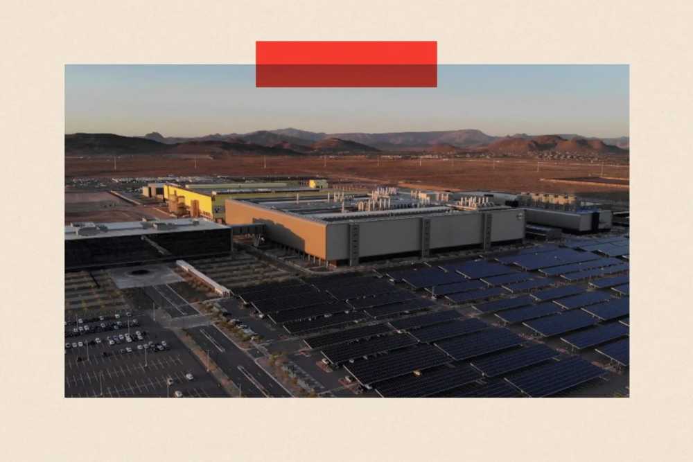 Large industrial facility with solar panels in front, surrounded by barren landscape and distant hills under a clear sky.