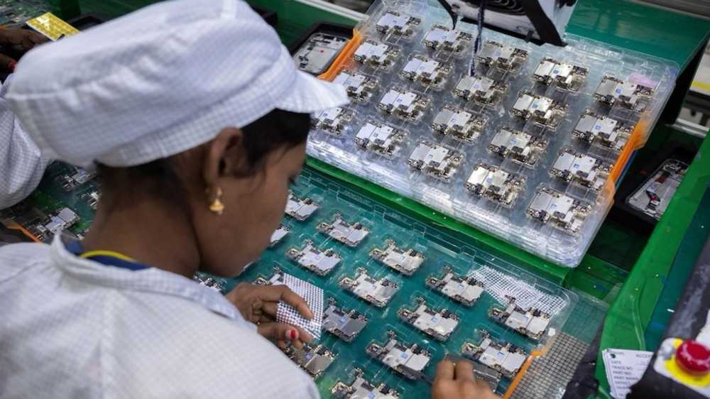 Worker assembling small electronic components at a factory, wearing a white cap and uniform, with trays of circuits on a green table in the work area.