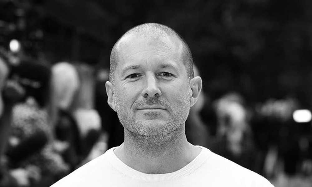 Jony Ive with a light stubble, wearing a plain shirt, stands in front of a blurred outdoor background.