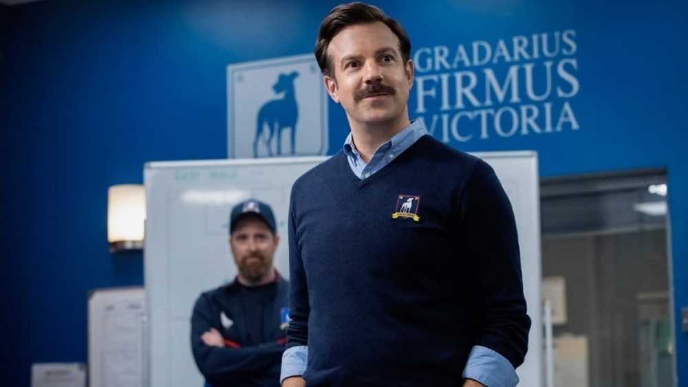Smiling man with mustache in blue sweater stands in front of whiteboard and blue wall, another man in a cap is blurred behind him.