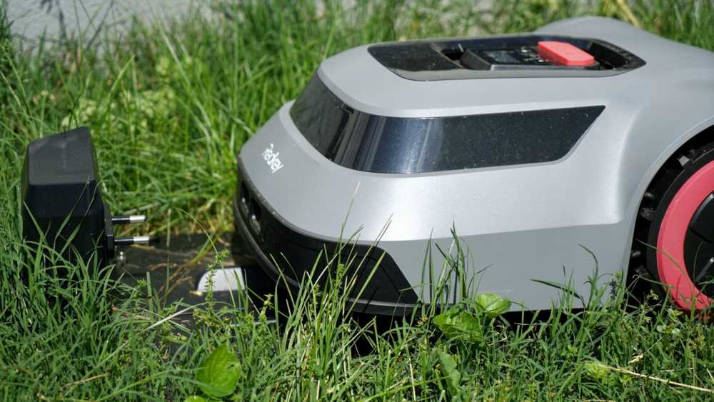 A robotic lawn mower in green grass approaches a charging station with a black plug.