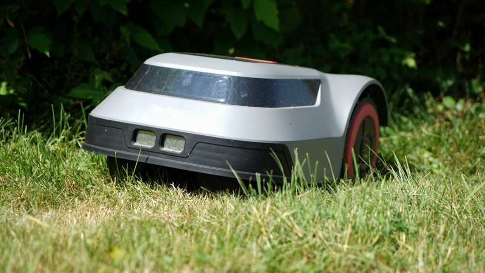 Gray robotic lawn mower on grass with a backdrop of green foliage.