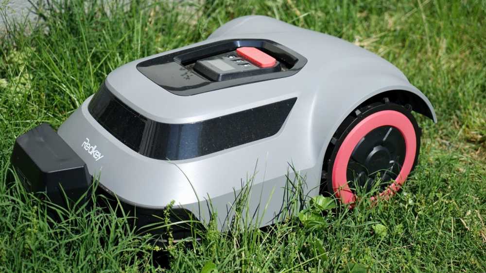 A silver robotic lawnmower with pink wheels sits on green grass, featuring a black panel on top with a red button.