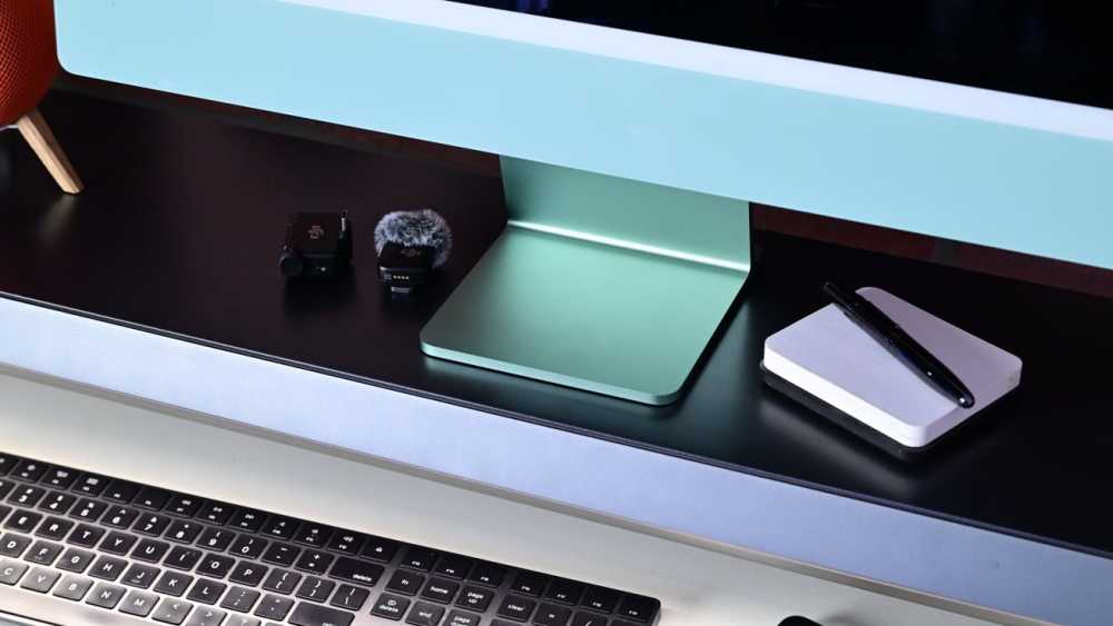 Desk setup with an iMac, wireless microphone, mini-laptop, and pen on a notebook.