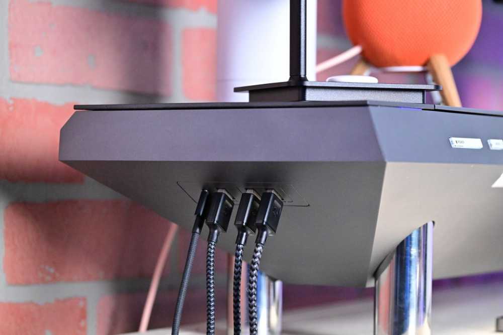 Black electronic device with multiple braided cables plugged in, set on a silver stand, with a brick wall background and a spherical orange object nearby.