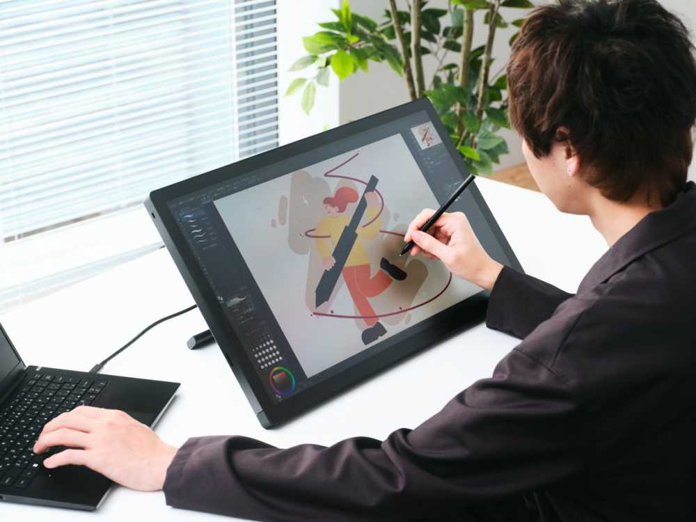 Person using a digital pen to draw colorful art on a large tablet, connected to a laptop, with a plant and window blinds in the background.