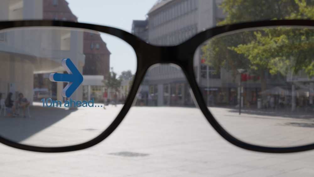 View through smart glasses displaying a digital arrow pointing forward with '10m ahead' text, set against an urban street background.