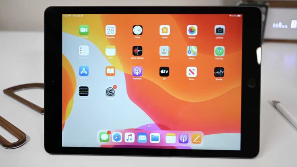 Tablet displaying various app icons on a screen with an orange and yellow wavy background, placed on a white surface with a stylus nearby.