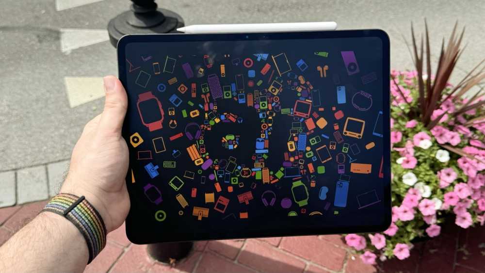 Hand holding a tablet displaying colorful icons forming 'WWDC' with a sidewalk and flowerbed in the background.