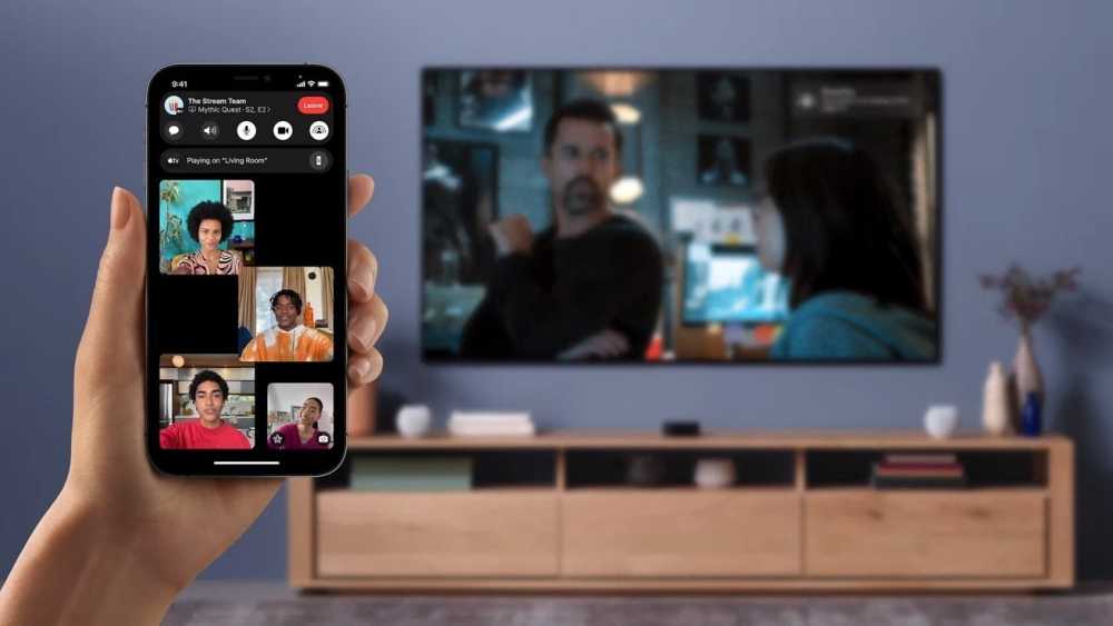 A hand holds a smartphone showing a video call with four people. A blurred TV in the background displays a scene with two characters.