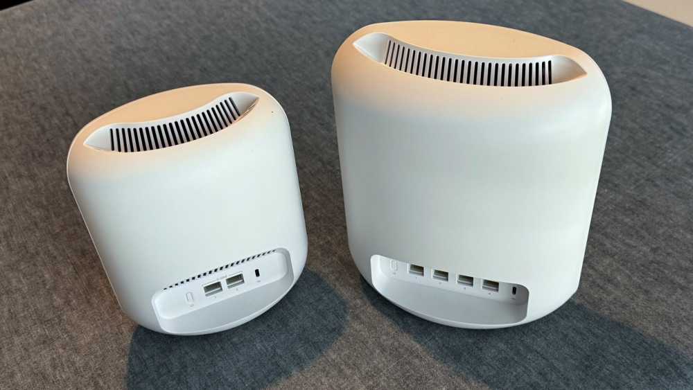 Two white, cylindrical electronic devices with ventilation slits on top and multiple ports on the back, placed side by side on a gray surface.