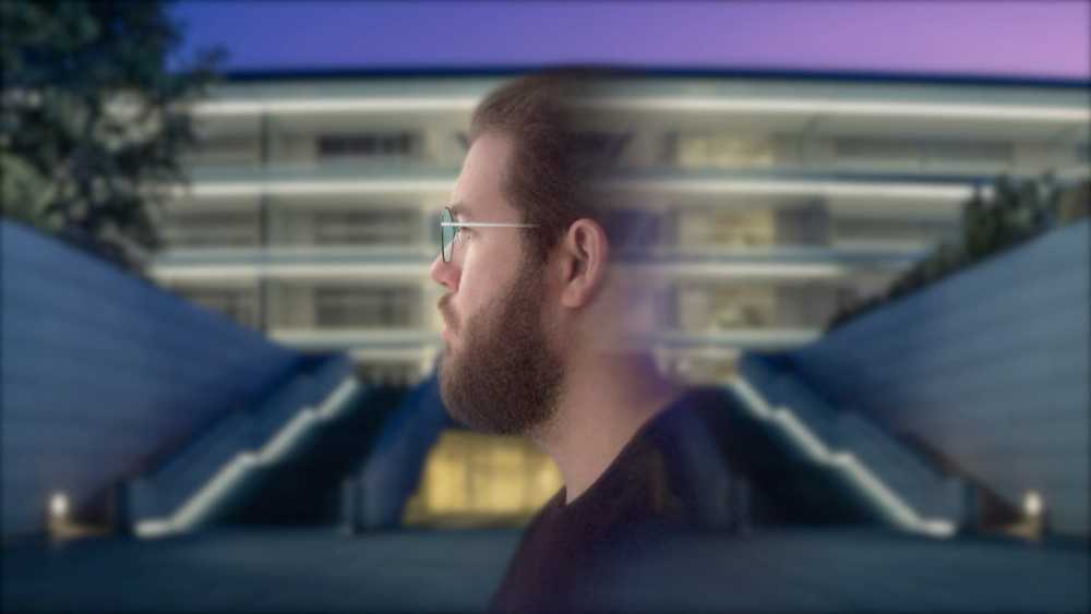 Side profile of a person with a beard and glasses, set against a blurred, modern, multi-story building background.