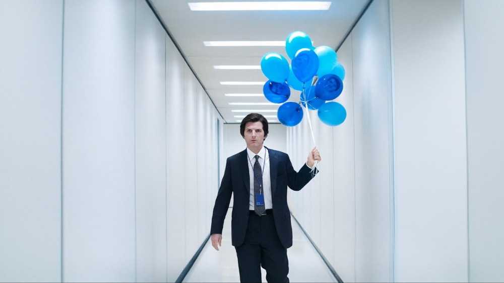 Man in suit walking down a narrow, brightly-lit hallway, holding a bunch of blue balloons.