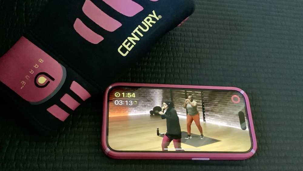 Smartphone displaying a workout video with two people exercising indoors. Nearby, a pink and black boxing glove rests on a textured surface.