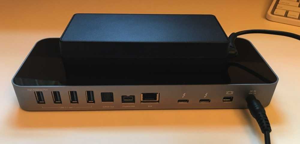A sleek electronic docking station with multiple ports, including USB, FireWire, and Ethernet. A black external power supply is resting on top, connected by a cable.