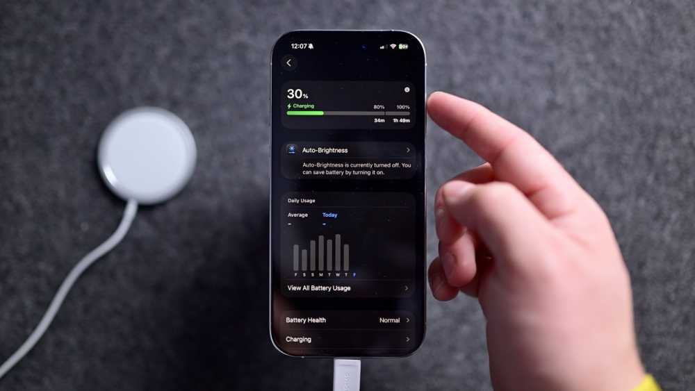 Hand holding a smartphone displaying battery charging status at 30%, alongside a wireless charging pad on a dark textured surface.