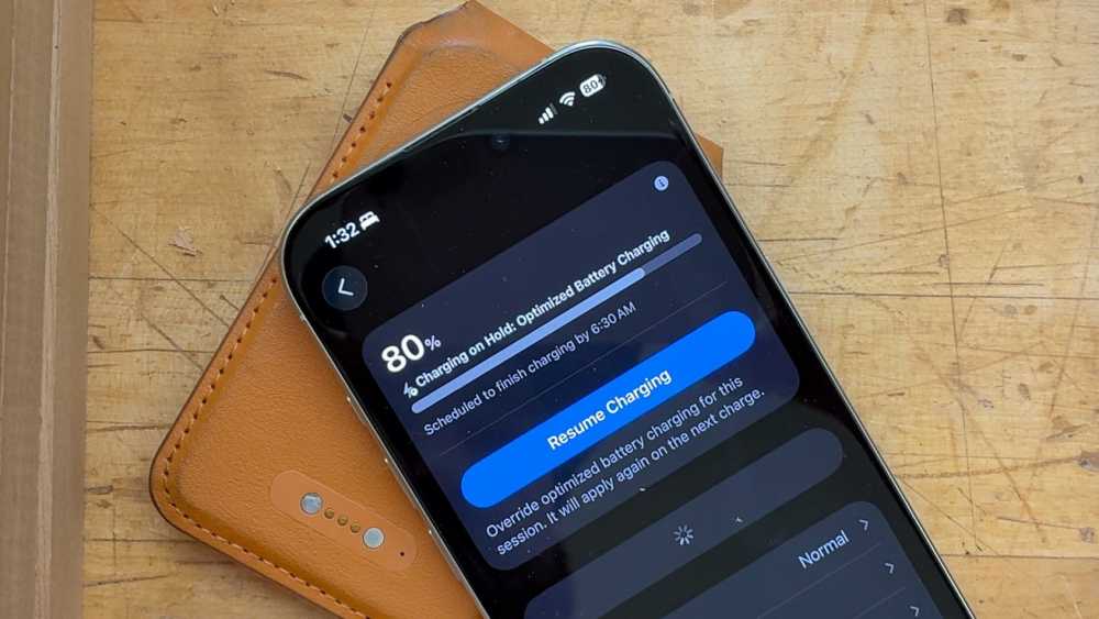 Smartphone on a wooden surface, showing battery status at 80 percent with optimized charging notification on the screen. Brown case partially visible underneath.