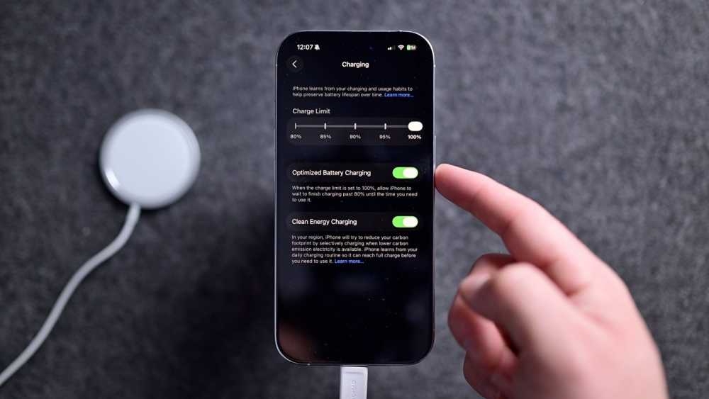 Smartphone charging settings screen with options for charge limit, optimized battery charging, and clean energy charging. A hand points towards the screen. Circular charging pad nearby.