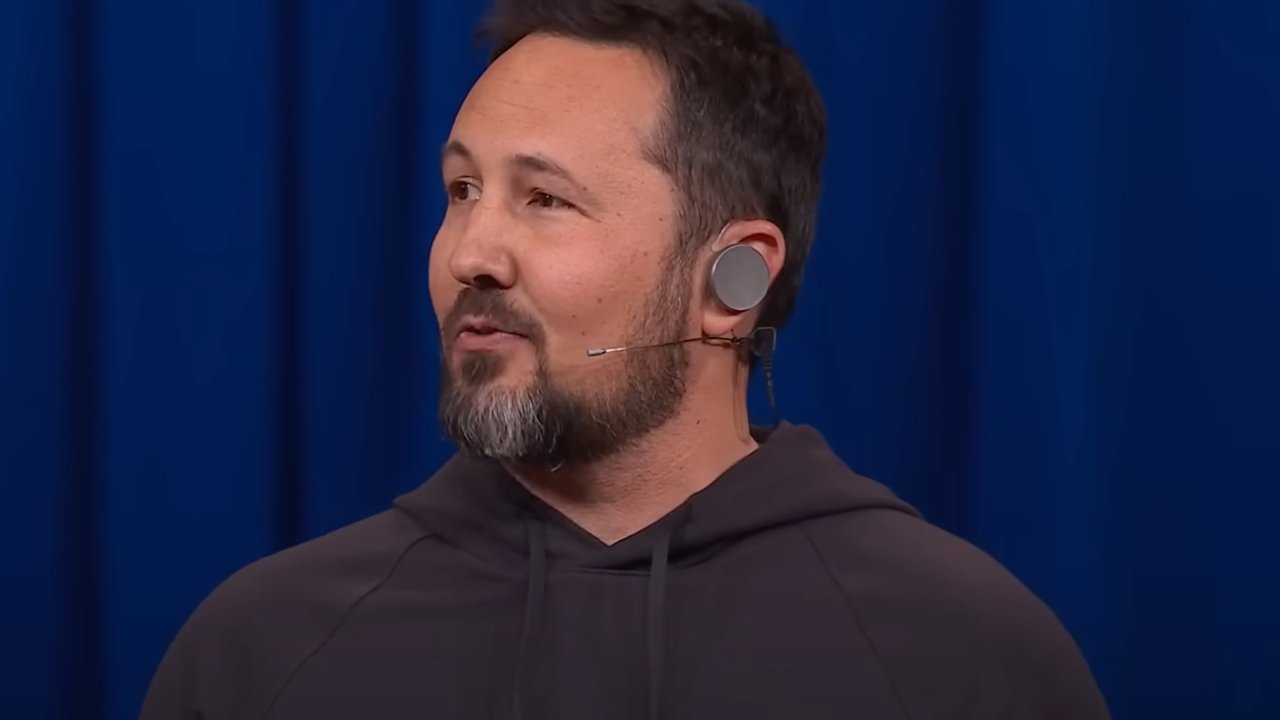 Man with short hair and beard wearing a hoodie and a 头显, speaking in front of a blue curtain.